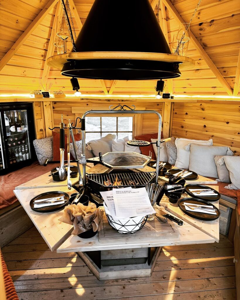 "Grill Haus Südsteiermark beim Landhaus Kainz von innen. Runder Tisch mit komfortablen Sitzbänken, in der Mitte Metall-Griller. Der perfekte Grillabend"