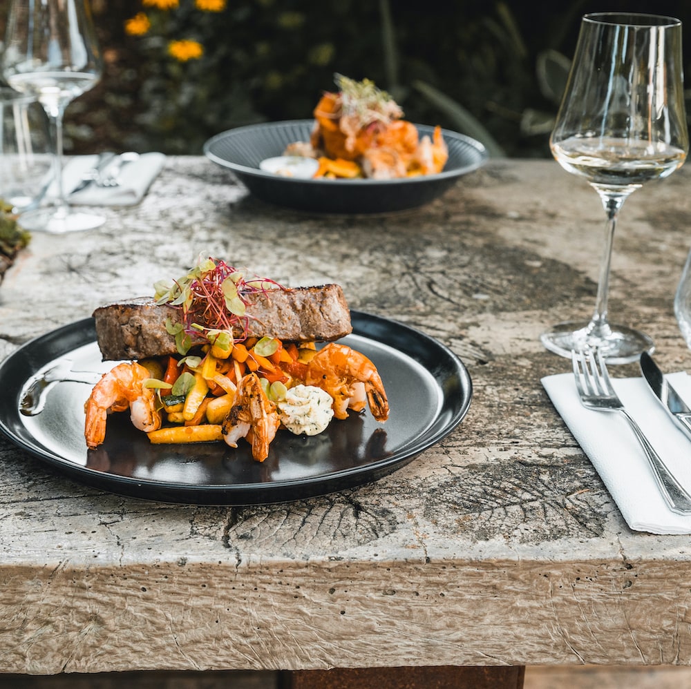 Speisekarte im Landhaus Kainz - edles Restaurant in der Südsteiermark. Steak mit Garnelen (Surf & Turf) auf Gemüse und Kräuterbutter, gedeckter Tisch im Garten mit Weingläsern – Speisekarte im Landhaus Kainz | Genuss & Wein aus der Südsteiermark